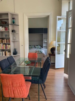 Un espace de vie compact comprenant une table à manger en verre, des chaises, une bibliothèque, un lit avec une couvre-lit à motifs et une porte donnant sur un espace extérieur.