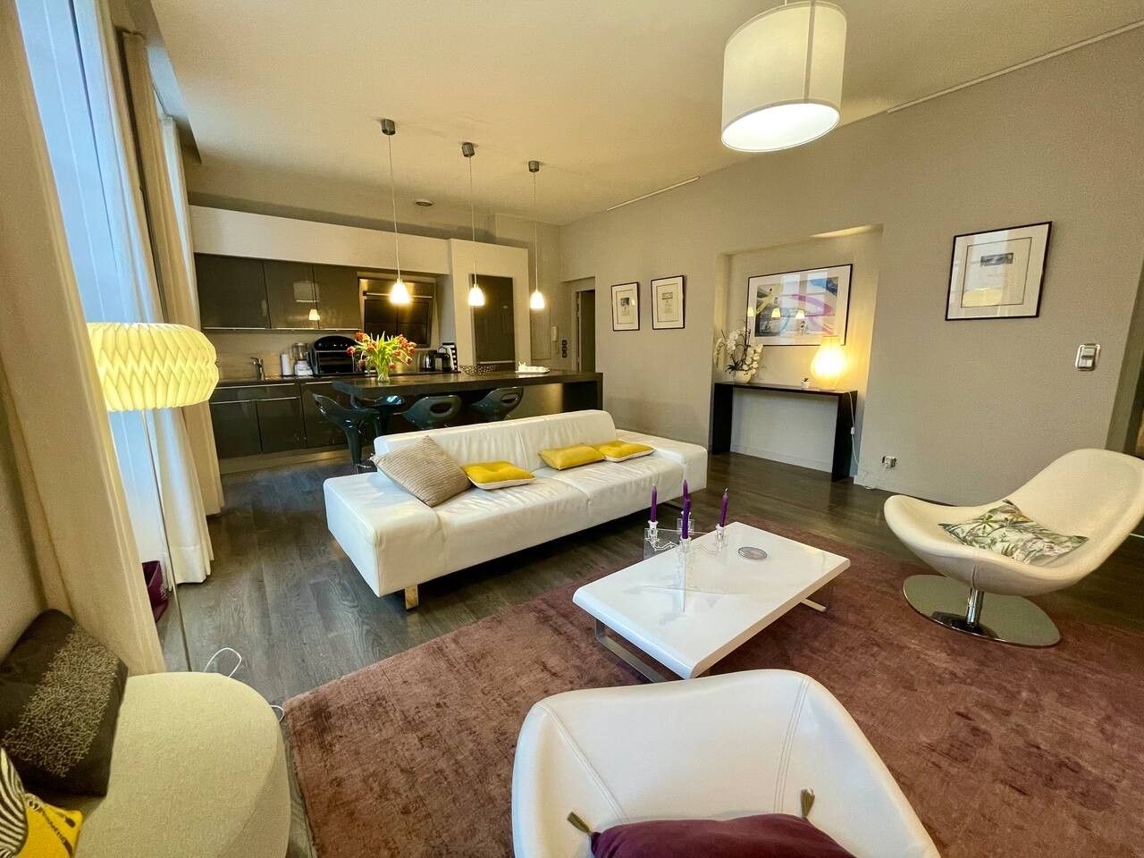 A modern living room with a white sofa, a coffee table, and a chair. There is a kitchen in the background with a dining table and chairs.