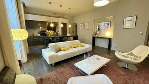 A modern living room with a white sofa, a coffee table, and a chair. There is a kitchen in the background with a dining table and chairs.