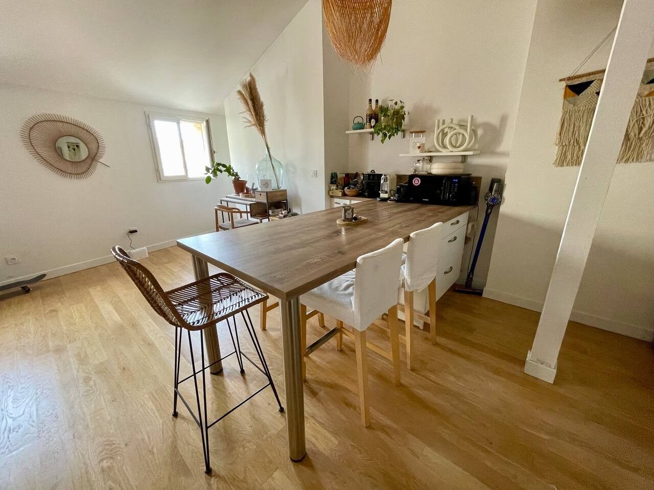 A dining area with a wooden table and chairs, a kitchen in the background, and a hanging basket.