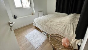 Een slaapkamer met een bed, een raam, een radiator, een wit vloerkleed en een rieten stoel.