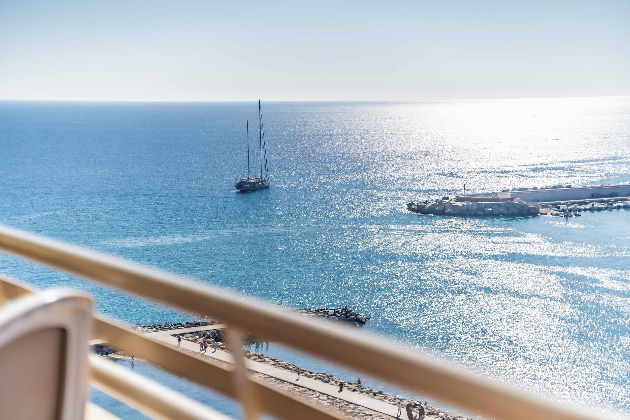 A balcony with a view of the sea, a sailboat, and a rocky breakwater.