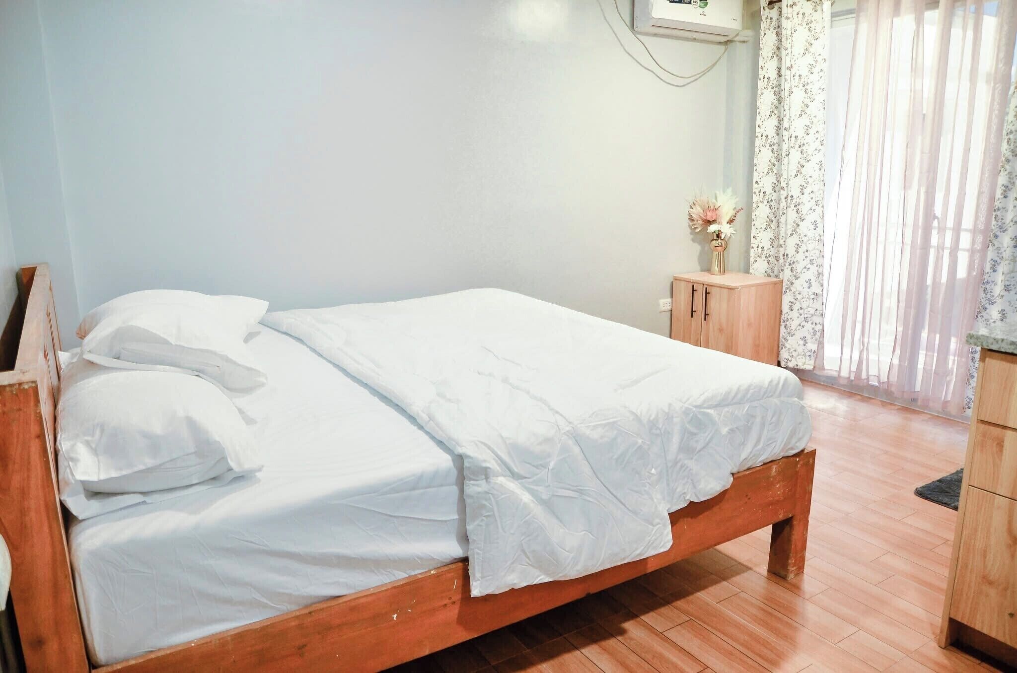 Una cama bien hecha con sábanas blancas en una habitación con suelo de madera, una mesita de noche de madera y un pequeño armario.