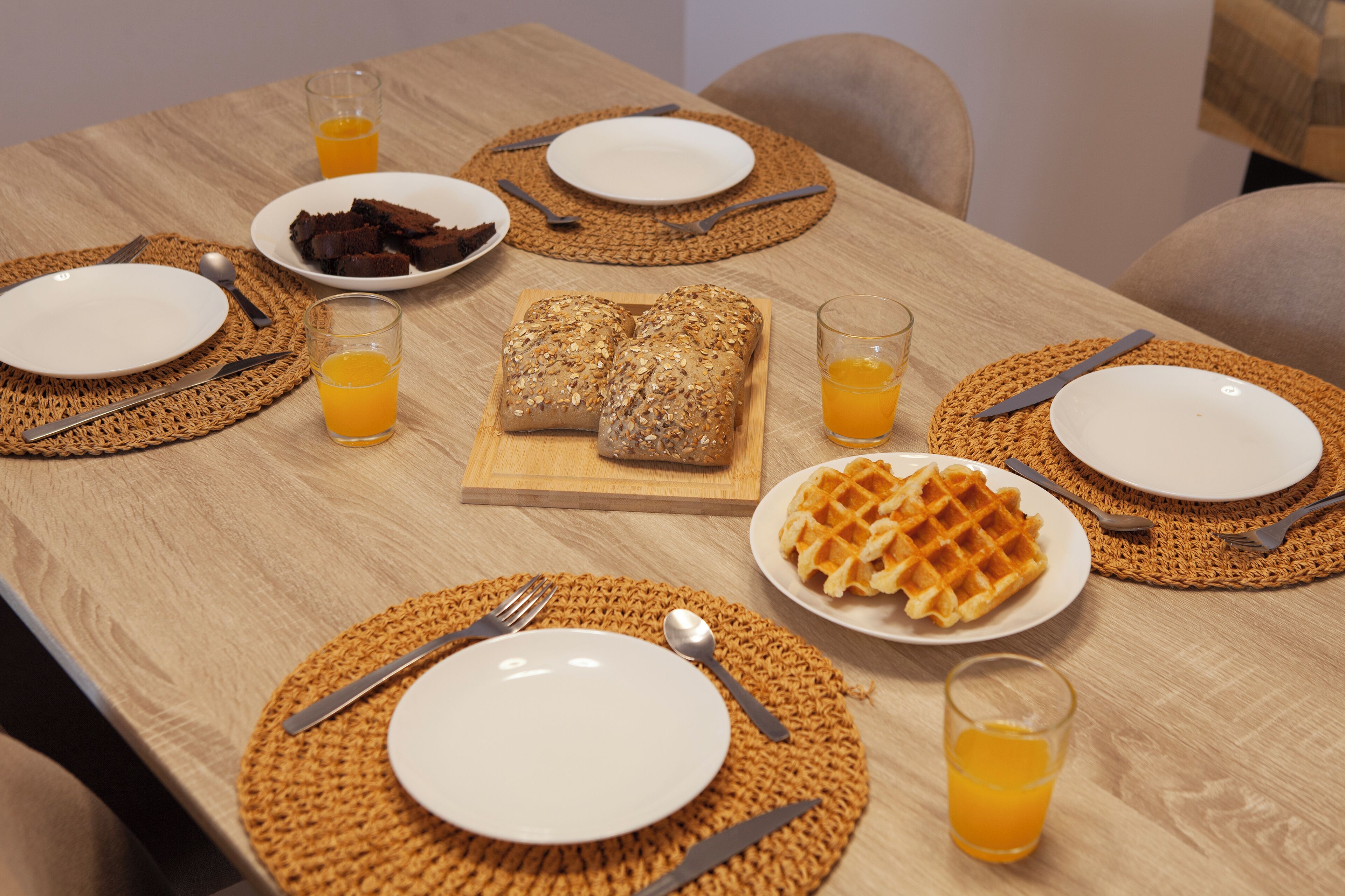 Una mesa de madera con un mantel tejido, platos, cubiertos y una hogaza de pan.