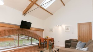 A spacious living room with a high ceiling, skylight windows, a flat-screen TV, a dining area with a table and chairs, and a cozy seating area with a sofa and cushions. - Strawberry line, flat 2 (Wells)
