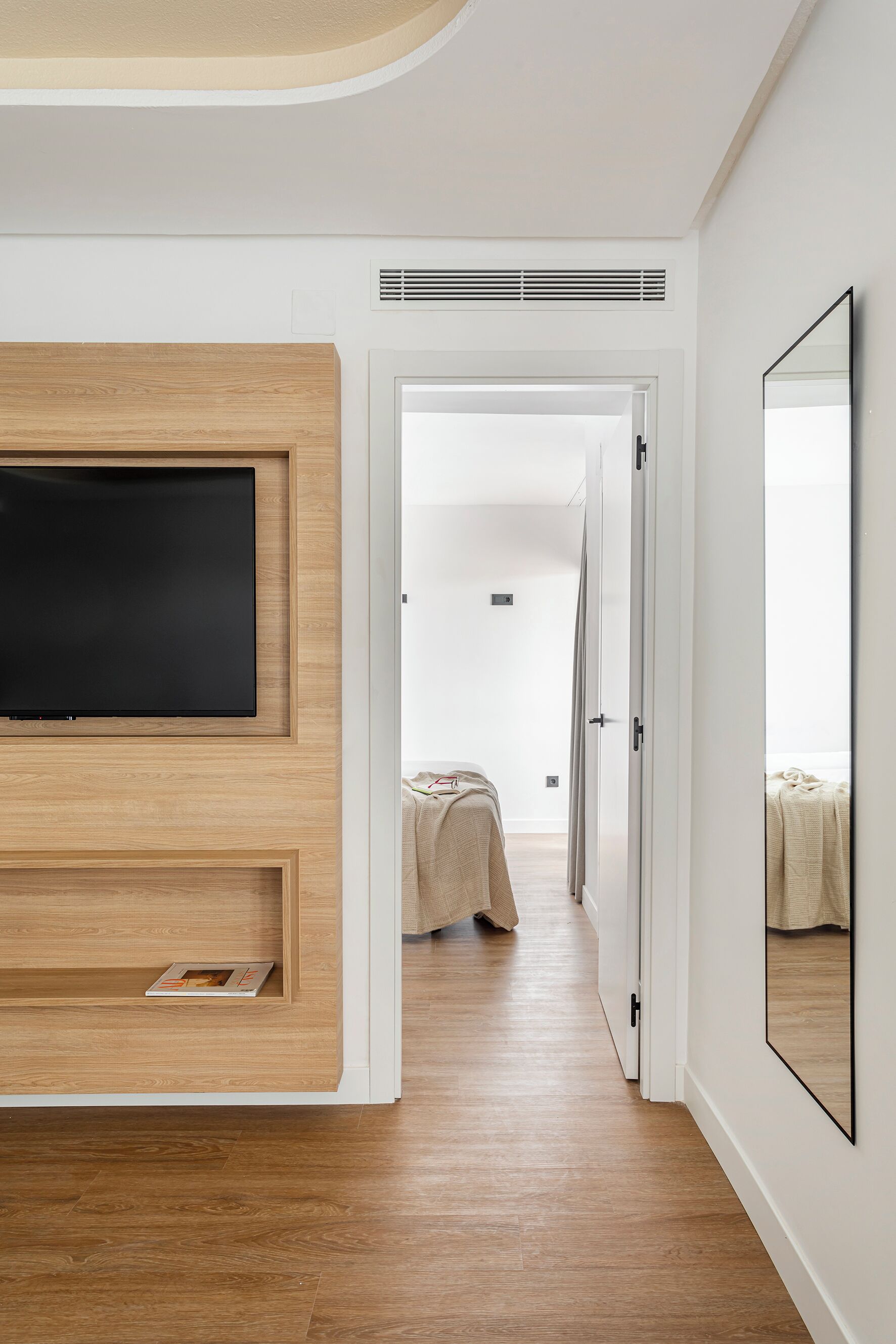 Una habitación de hotel moderna con un televisor de pantalla plana grande montado sobre un panel de madera, un espejo grande en la pared y suelo de madera.
