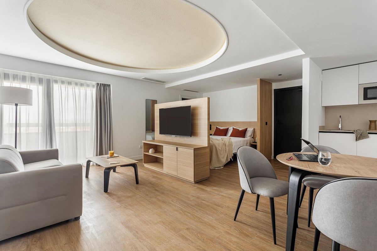 A modern hotel room with a flat-screen TV, a sofa, a dining table, and a bed with a wooden headboard.