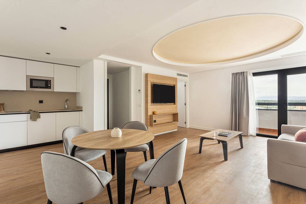 A modern hotel room with a dining area, a kitchen, a large window with a view, and a ceiling light fixture.