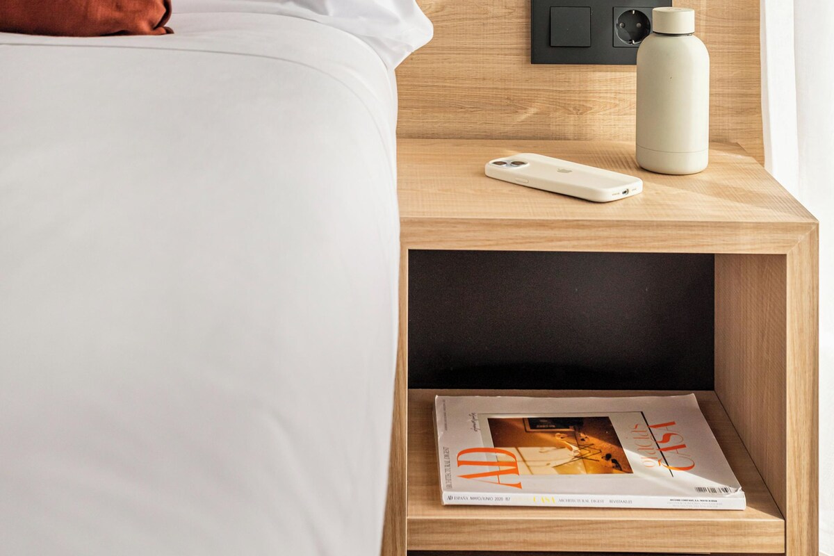 Una cama bien tendida con almohadas blancas y marrones, una mesita de noche con una lámpara, una botella de agua y una revista.