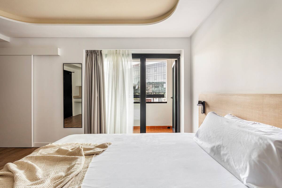 A modern hotel room with a large bed, a view of the city through a sliding door, and a minimalist design.