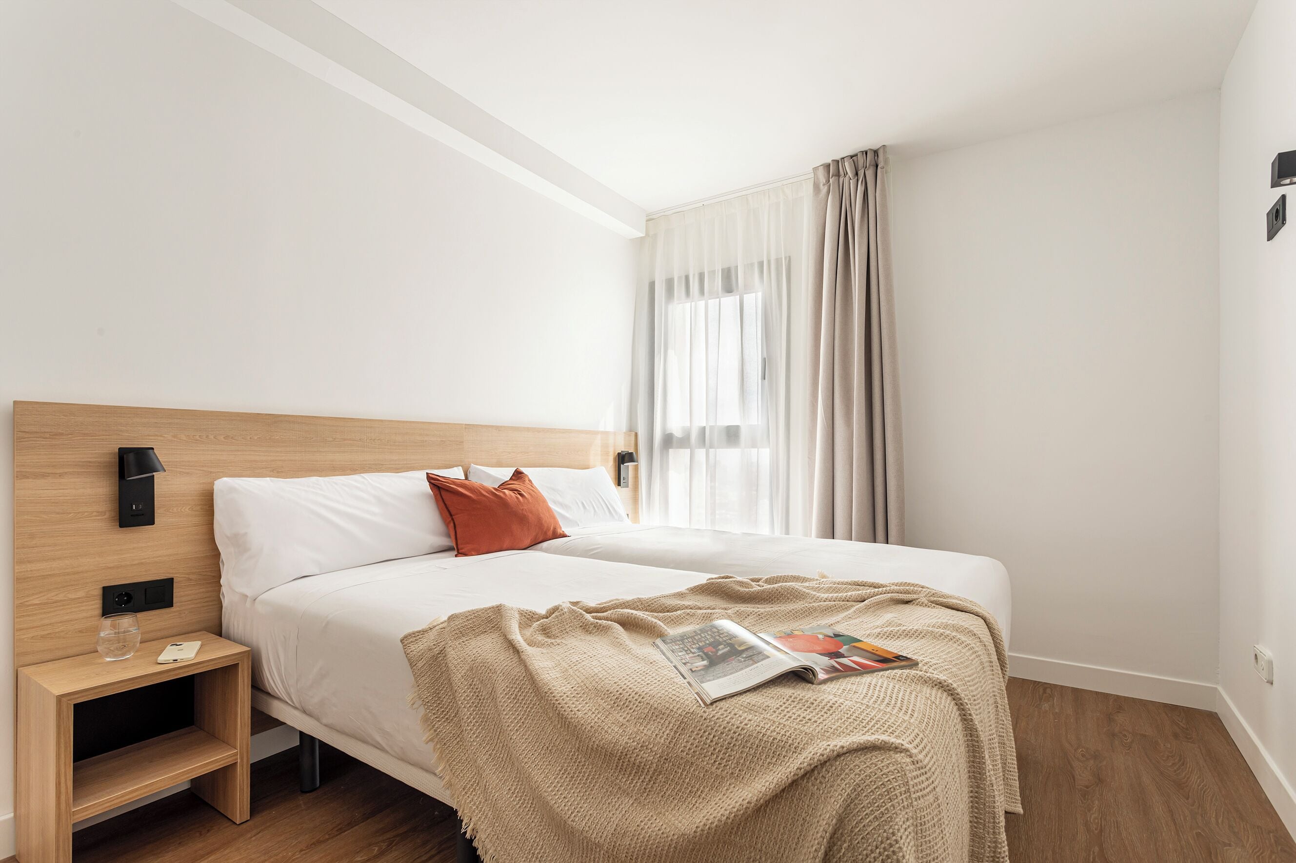 A modern hotel room with a wooden headboard, a single bed with white bedding, a beige throw blanket, and a magazine on the bed.