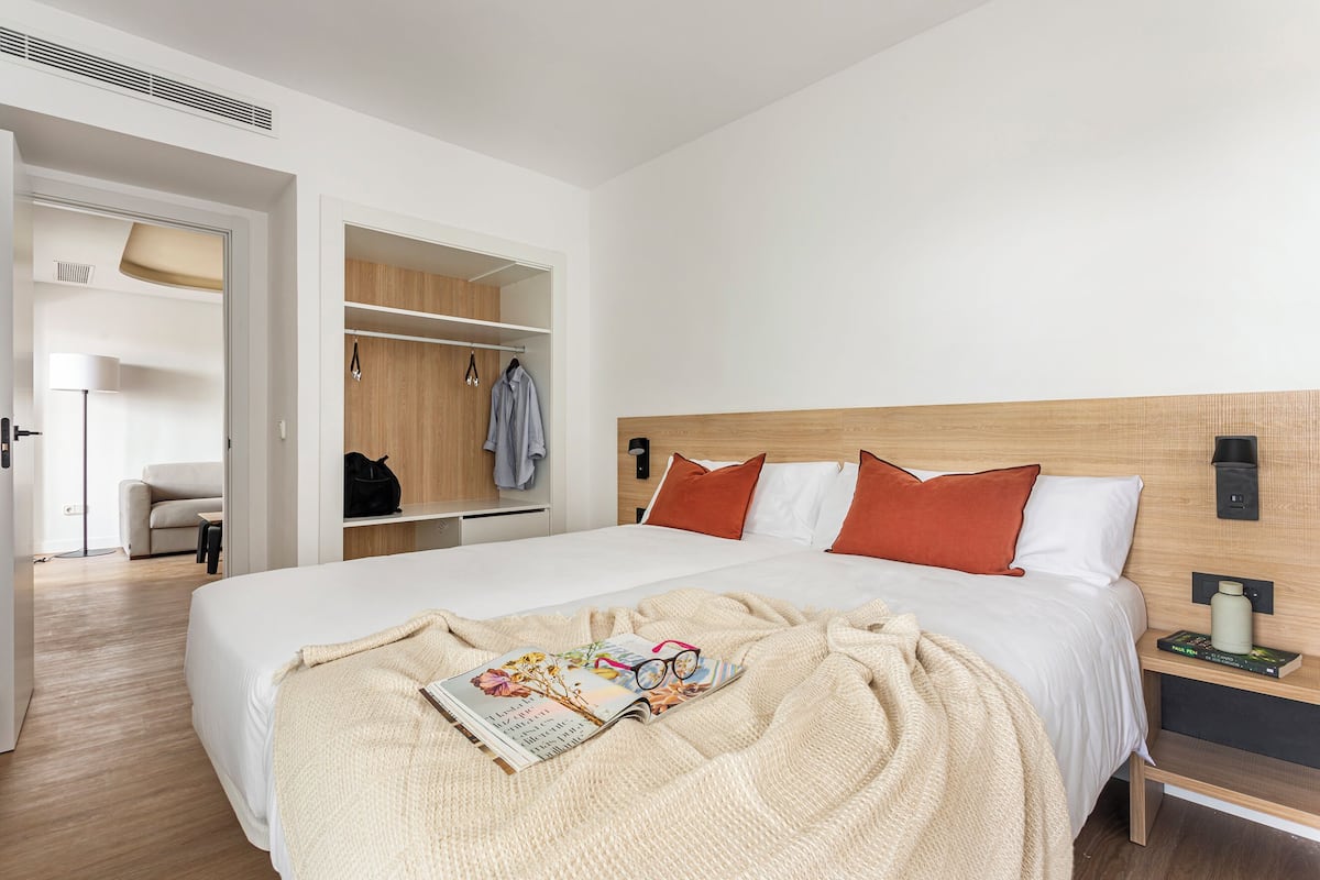 A modern hotel room with a large bed, a bedside table with a book, a wardrobe, and a seating area with a chair and lamp.