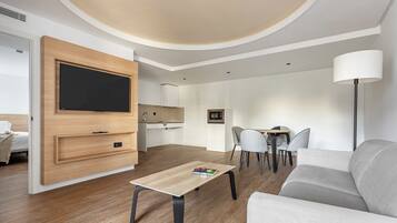 Una habitación de hotel moderna con un televisor de pantalla plana montado en un panel de madera, una zona de comedor con una pequeña mesa y sillas, un sofá y una lámpara de pie.