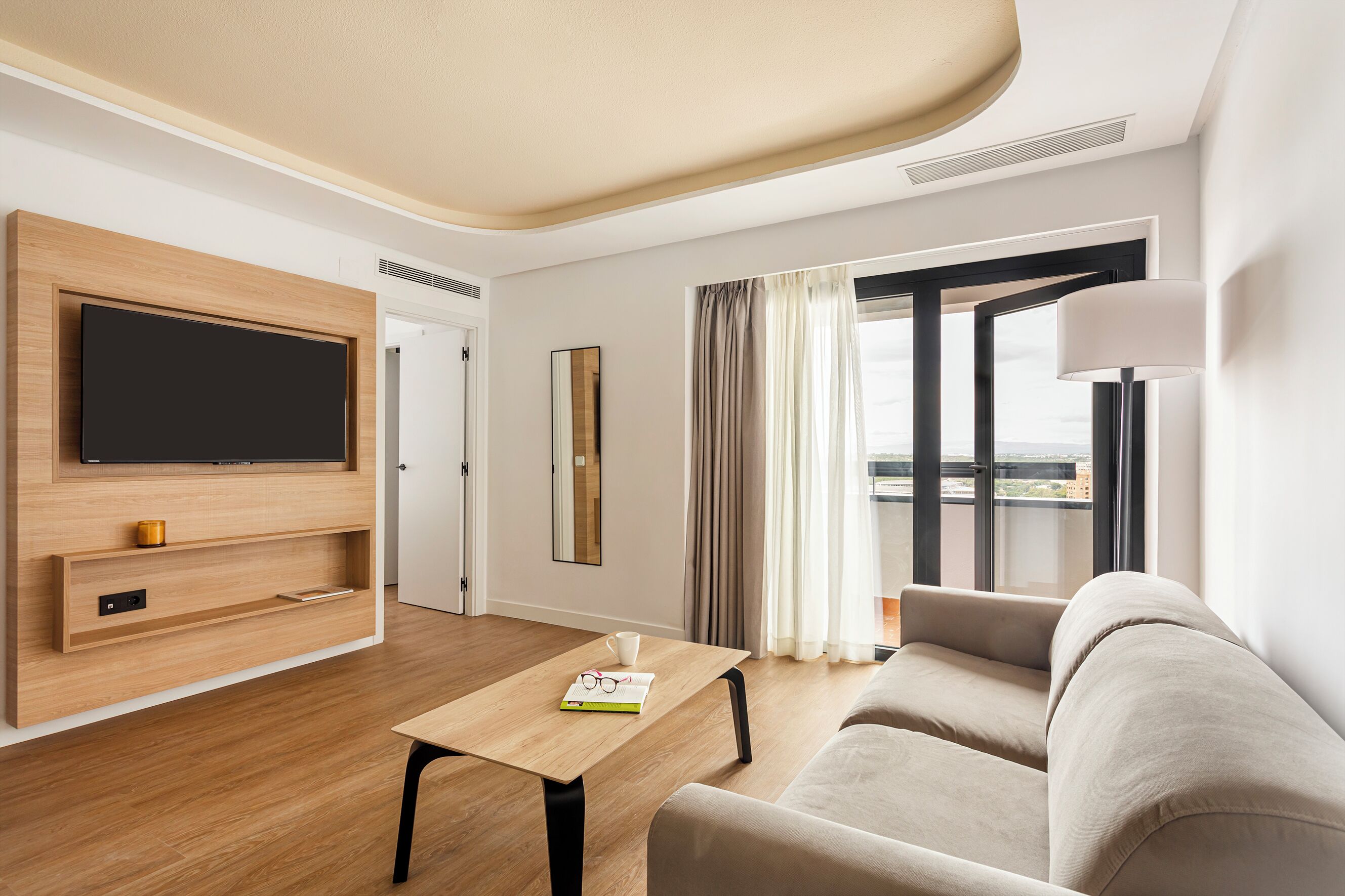 A modern hotel room with a flat-screen TV mounted on a wooden wall, a sofa, a coffee table, and a large window with curtains.