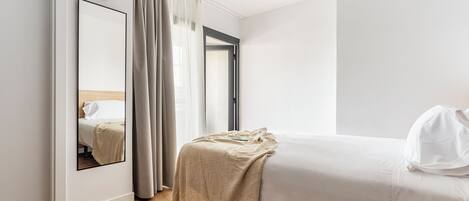 A hotel room with a bed, a mirror, a window with curtains, and a wooden floor.