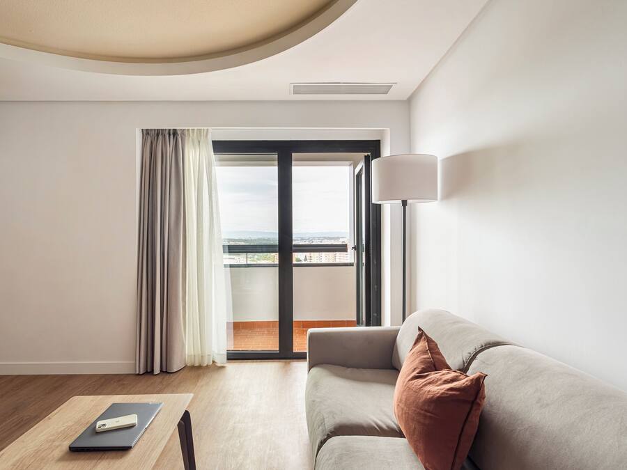 A modern hotel room with a sofa, a table with a laptop, and a view of the outdoors through a large window.
