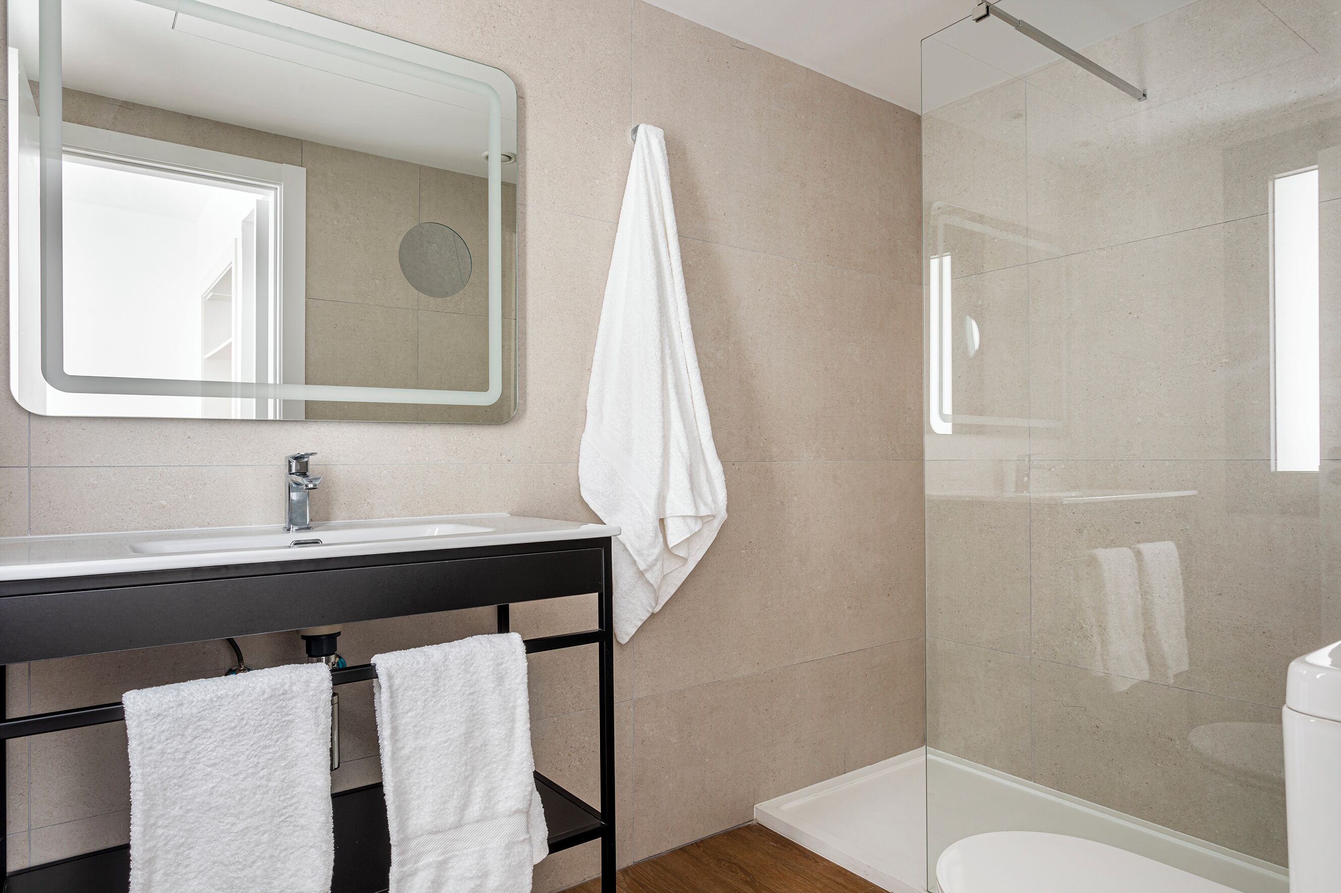 A modern bathroom with a large mirror, a sink, a black shelf with towels, and a glass shower enclosure.