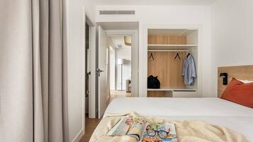 A modern hotel room with a bed, a bedside table with a magazine and glasses, a wardrobe, and a nightstand with a lamp.