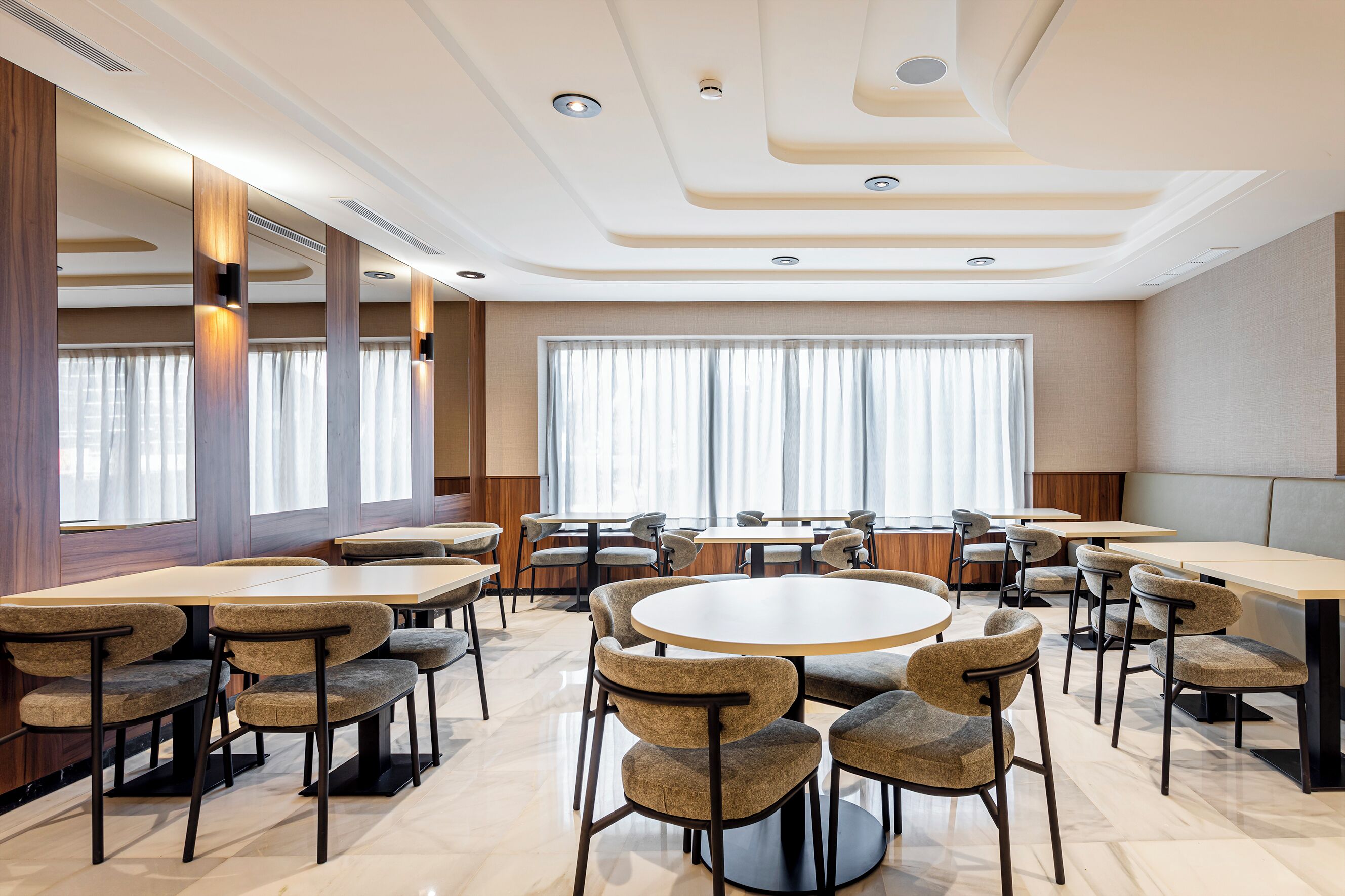 Un'area pranzo moderna con tavoli rotondi e sedie imbottite, ampie finestre con tende leggere e un soffitto con illuminazione a incasso.