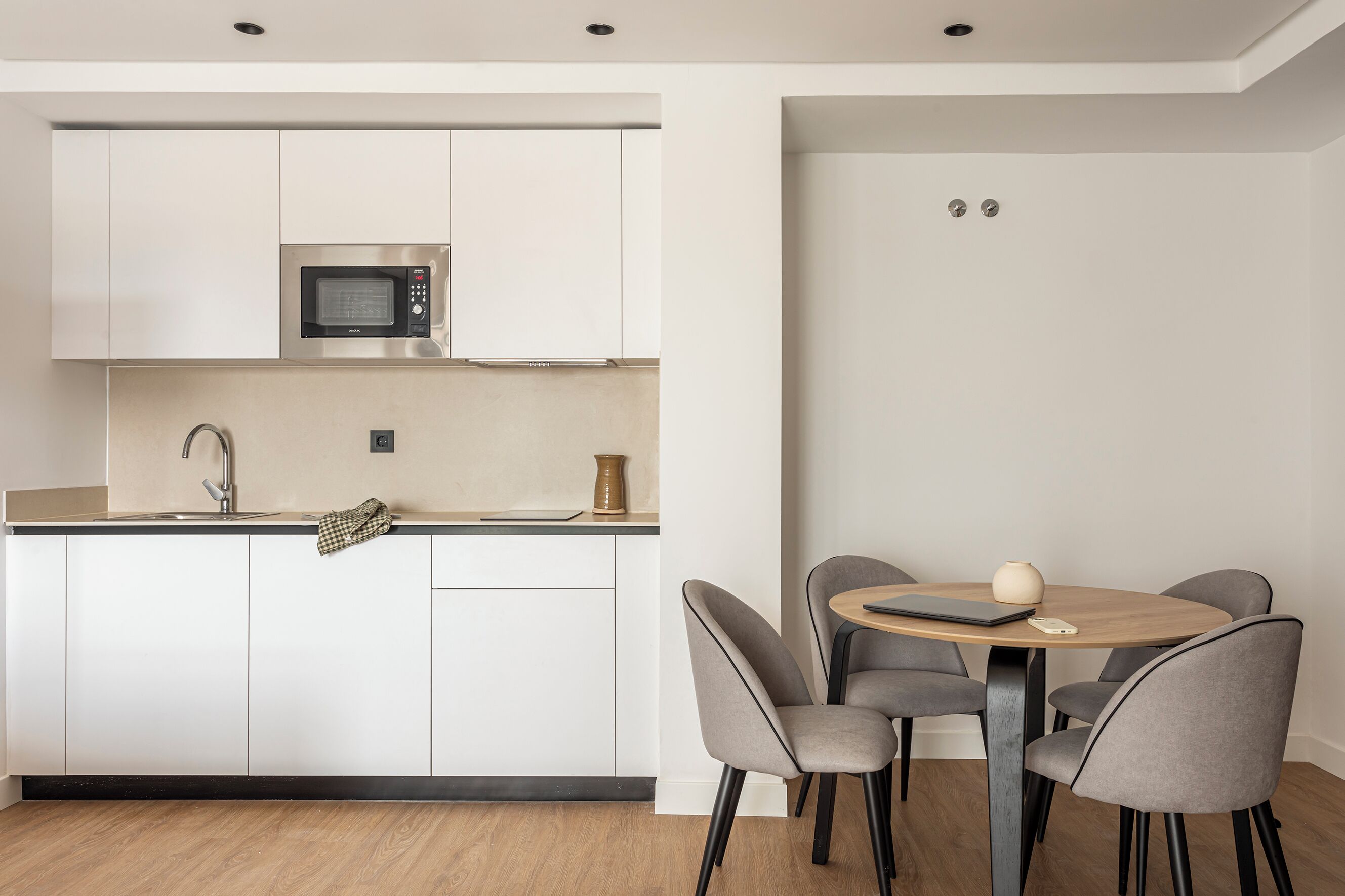 Una cocina moderna con armarios blancos, un microondas integrado y una mesa redonda de comedor con cuatro sillas grises.