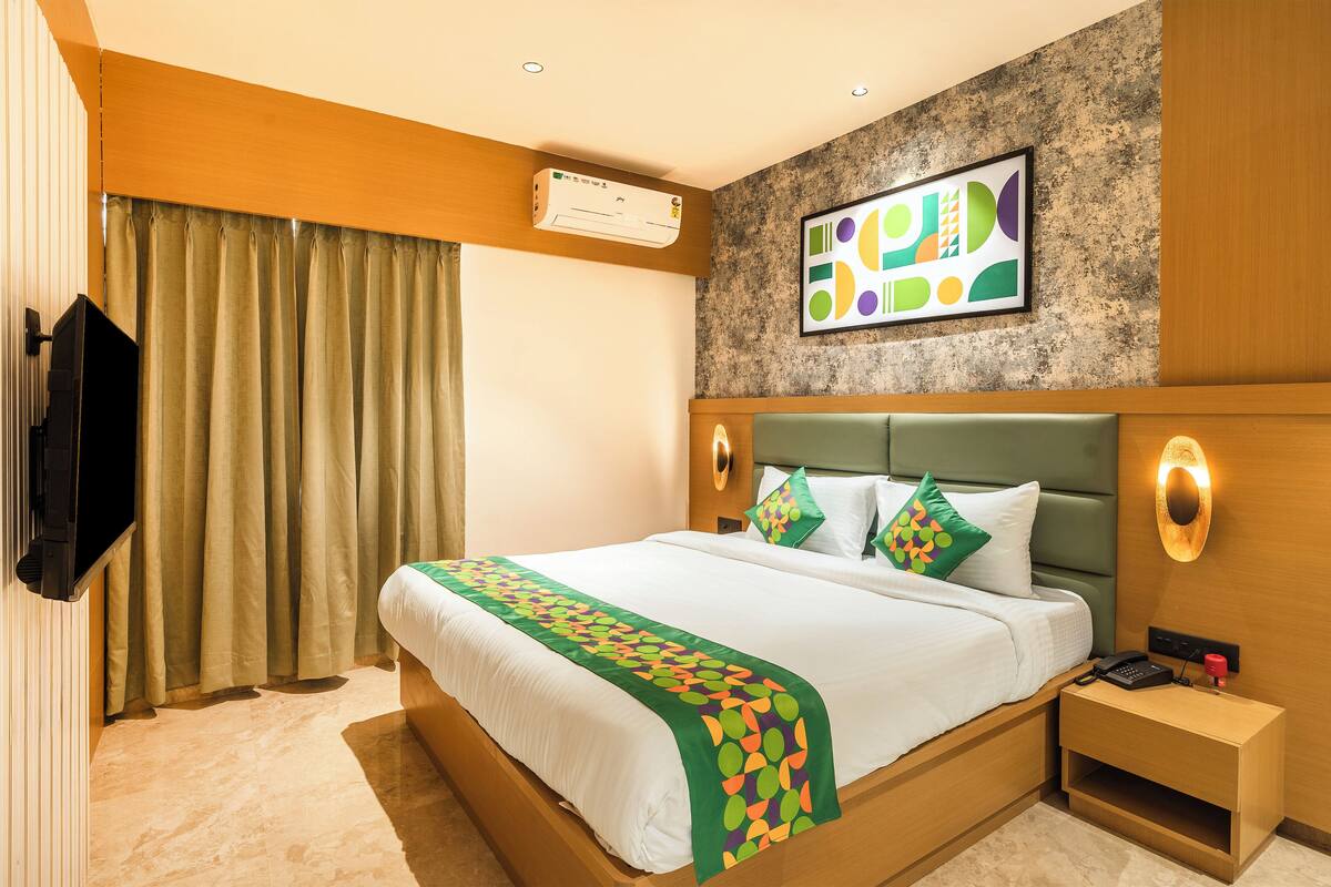 A hotel room with a bed, a flat-screen TV mounted on the wall, a bedside table with a lamp, and a wall-mounted air conditioner.