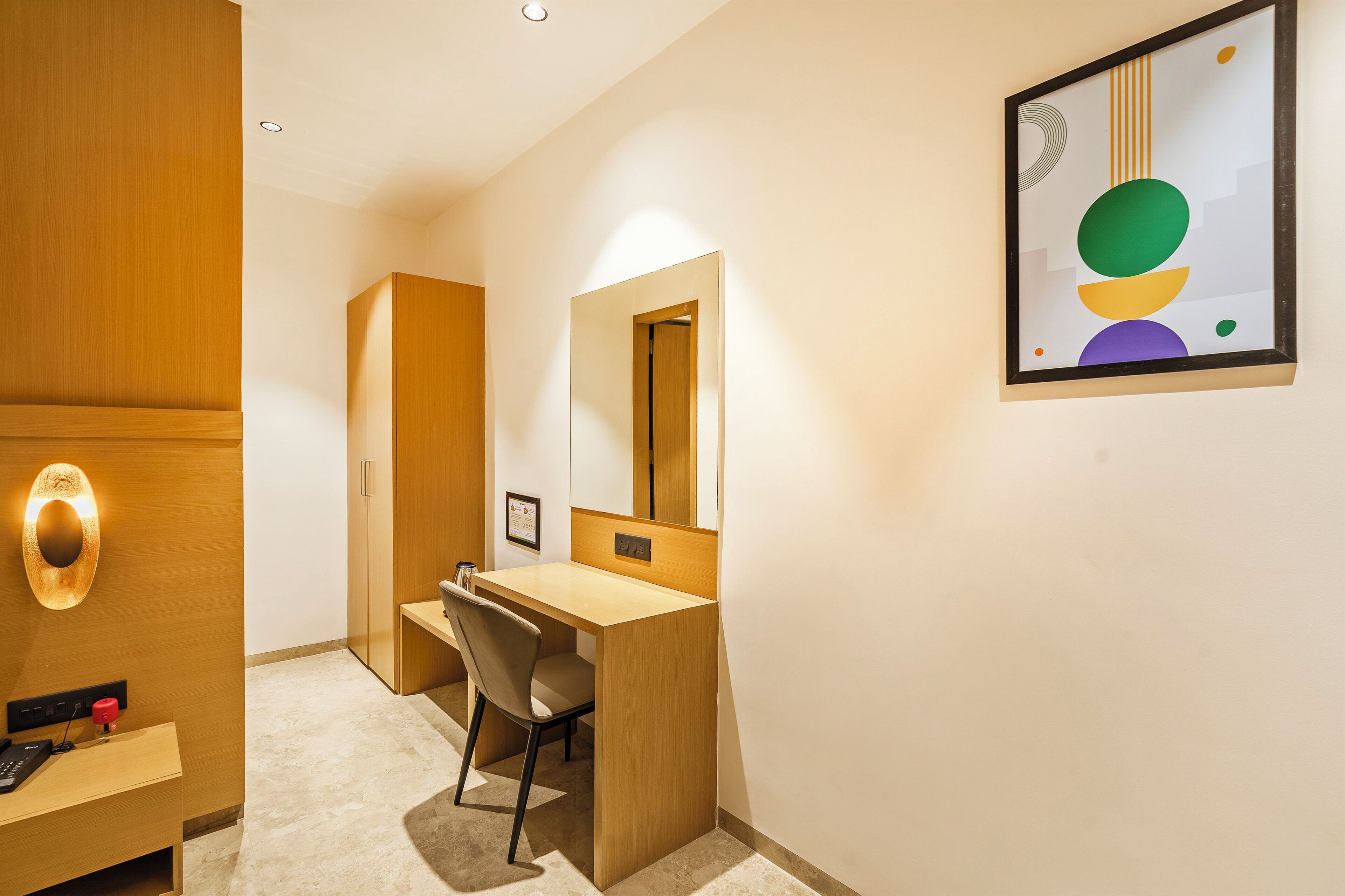A hotel room with a wooden desk, a chair, a large mirror, and a framed abstract artwork on the wall.