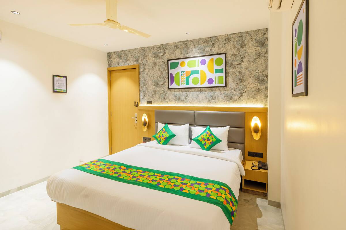 A hotel room with a bed, a headboard with wall-mounted lights, a ceiling fan, and a framed abstract artwork on the wall.