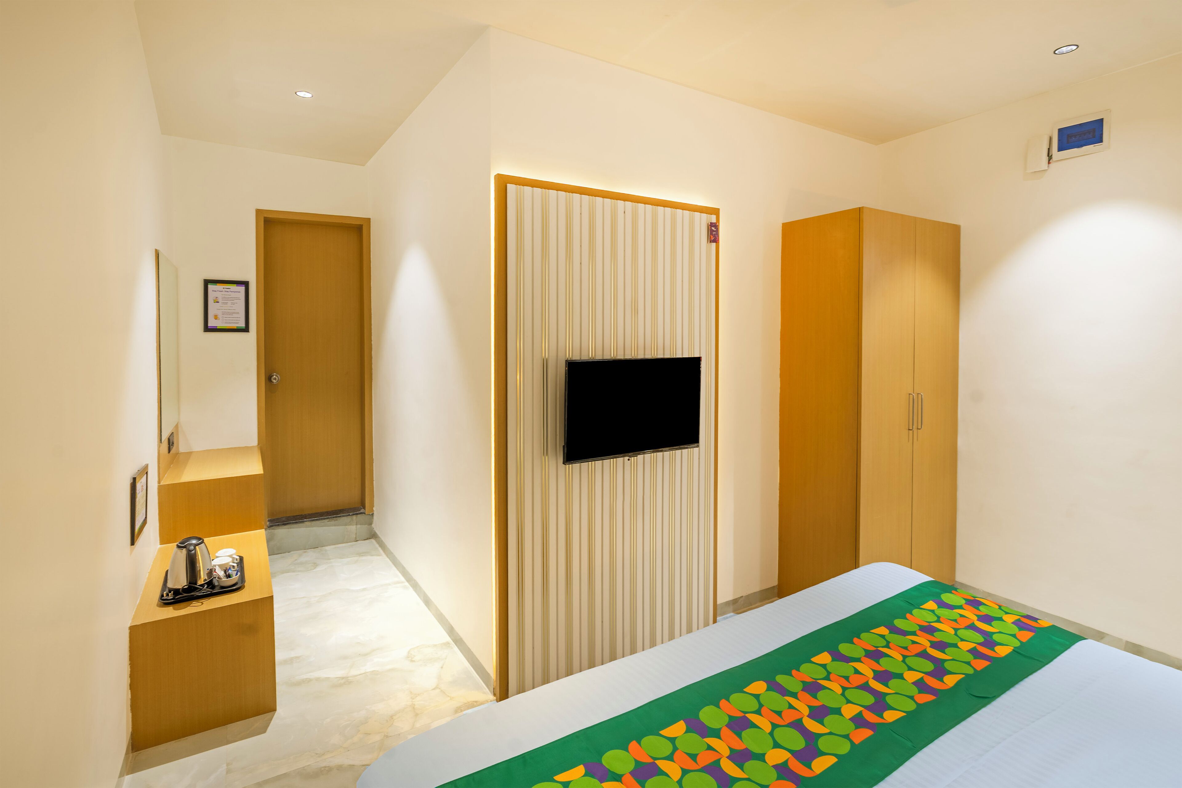 A hotel room with a bed, a television mounted on the wall, a wardrobe, and a small seating area with a coffee maker.