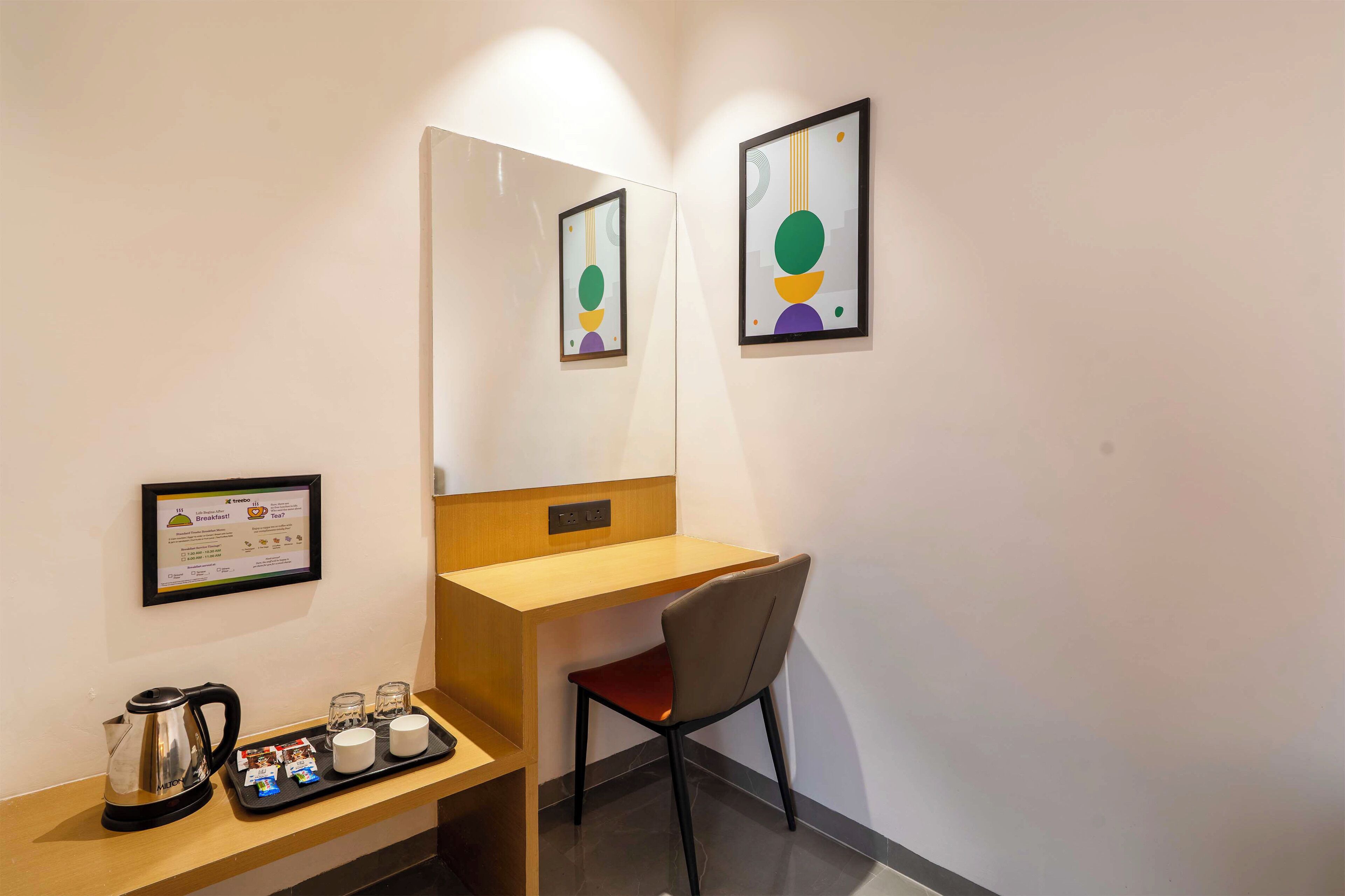A hotel room with a wooden desk, a chair, a coffee maker, and a tray with a mug and a small box. There are framed pictures on the wall and a framed certificate on the wall.