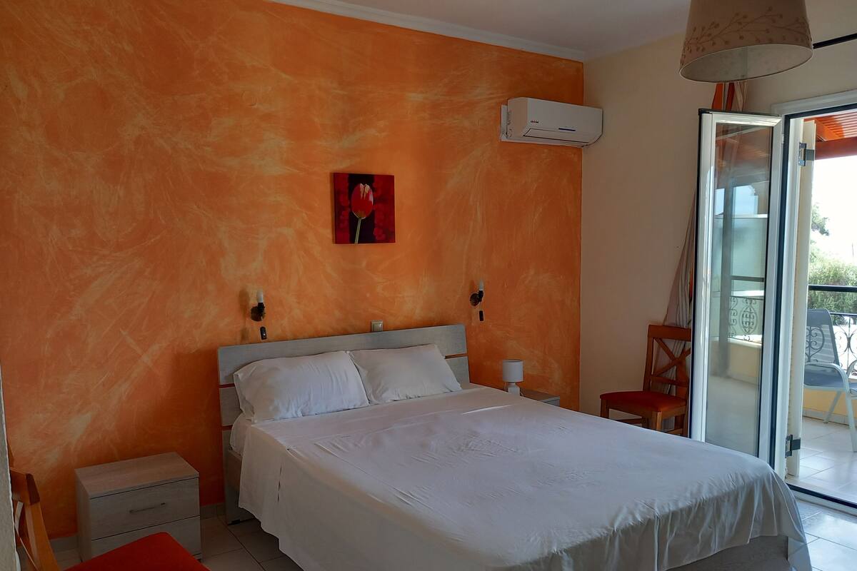 A bedroom with a bed, a nightstand, a chair, a red cushion, an air conditioning unit, and a window with a view.