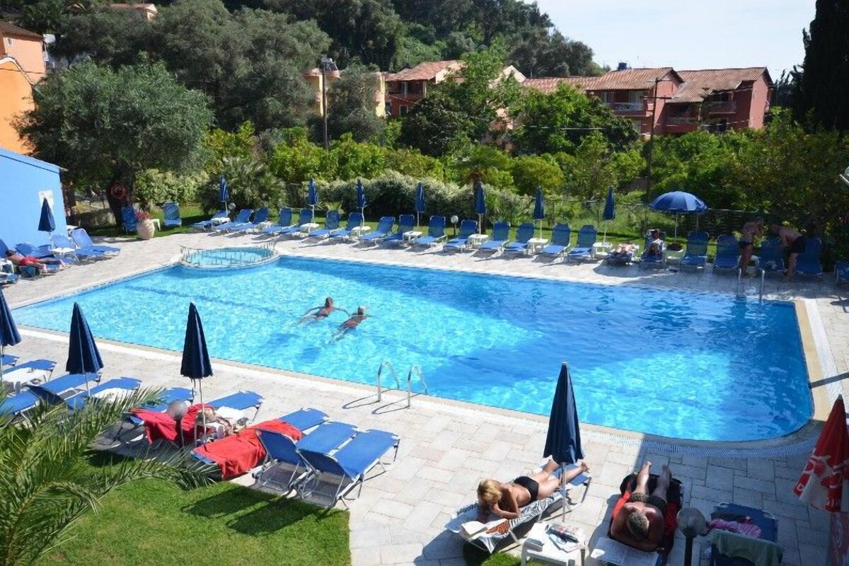 A swimming pool with lounge chairs and umbrellas, surrounded by greenery and buildings.