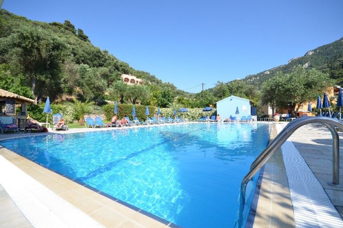 A swimming pool with lounge chairs and umbrellas, surrounded by greenery and hills.