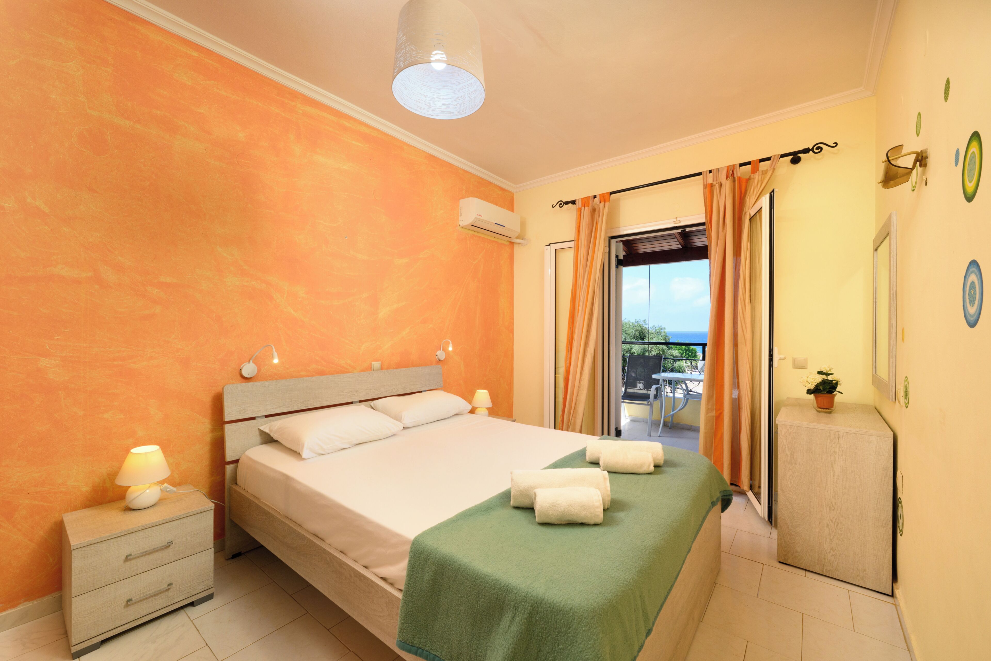 A bedroom with a bed, bedside table, lamp, and a view of the outside.