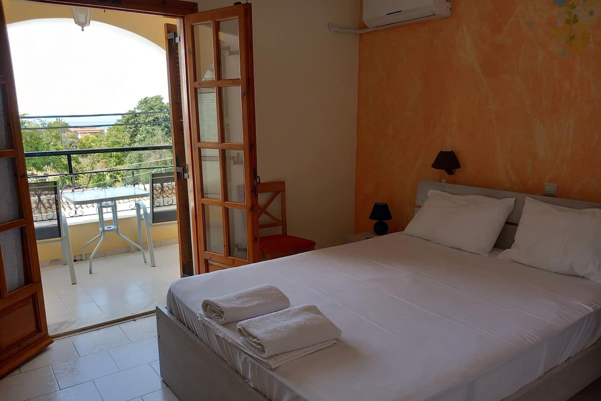 A hotel room with a bed, a nightstand, a lamp, a balcony with a table and chairs, and an open door leading outside.