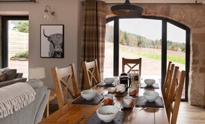 A dining area with a wooden table set for four, chairs, a framed artwork, and a view of a rural landscape.