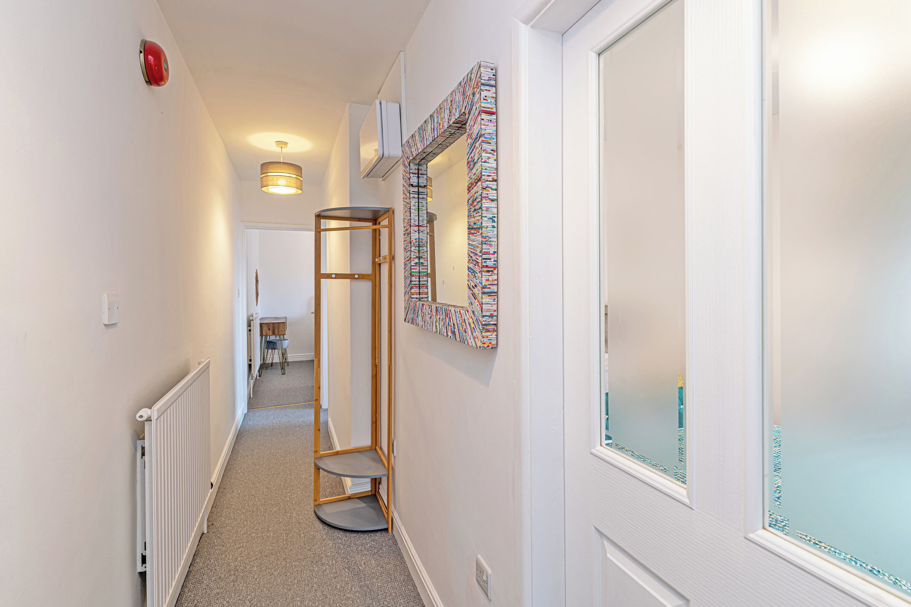 A hallway with a radiator, a wooden chair, a small table, and a mirror with a colorful frame.