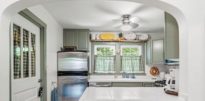 A kitchen with a stainless steel refrigerator, a built-in oven, a ceiling fan, and a window with checkered curtains.