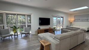 A spacious living room with a large sectional sofa, a wooden coffee table, and two armchairs. There are large windows offering a view of the outdoors.