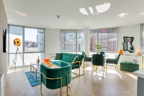 Un salon moderne avec un canapé d’angle, une table à manger et des chaises. Une grande fenêtre laisse entrer la lumière naturelle et offre une vue sur les bâtiments environnants.