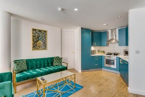 A modern living room with a green sofa, a glass coffee table, and a blue rug. Adjacent is a kitchen with teal cabinets, a stainless steel oven, and a white backsplash. - 404 Hatton Garden Family Flat - Free Secure Parking (Liverpool)