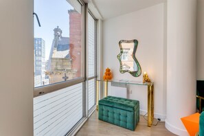 A modern room with a large window, a green ottoman, a glass console table with decorative items, and a mirror on the wall.