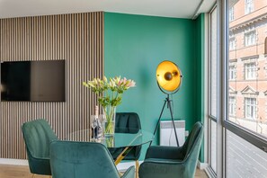 A modern room with a green wall, a flat-screen TV, a glass table with chairs, a tripod lamp, and a window with a view of buildings. - 404 Hatton Garden Family Flat - Free Secure Parking (Liverpool)