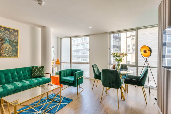 A modern living room with a green sofa, a glass coffee table, and a dining area with green chairs and a round table.