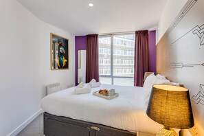 Una habitación de hotel con una cama grande, una ventana con cortinas, una obra de arte colgada en la pared, una lámpara de noche y una bandeja con artículos de desayuno.