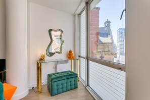 Une pièce moderne avec une console en verre, un pouf vert, un miroir décoratif et une vue sur le paysage urbain à travers de grandes fenêtres.