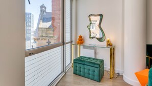 A modern room with a large window, a green ottoman, a glass console table with decorative items, and a mirror on the wall.