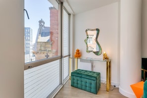 Une pièce moderne avec une grande fenêtre, un pouf vert, une console en verre agrémentée d’objets décoratifs et un miroir au mur.