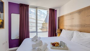 A hotel room with a large bed, a window with curtains, a headboard with a geometric design, and a tray with croissants and coffee cups.