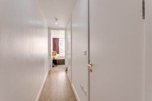 A narrow hallway with white walls and wooden flooring leading to a bedroom with a bed and a nightstand. - 202 Hatton Garden 2-Bed Family Flat with Secure Parking (Liverpool)