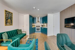 A modern living room with a teal sofa, a glass coffee table, and a TV mounted on a wooden panel wall. - 202 Hatton Garden 2-Bed Family Flat with Secure Parking (Liverpool)
